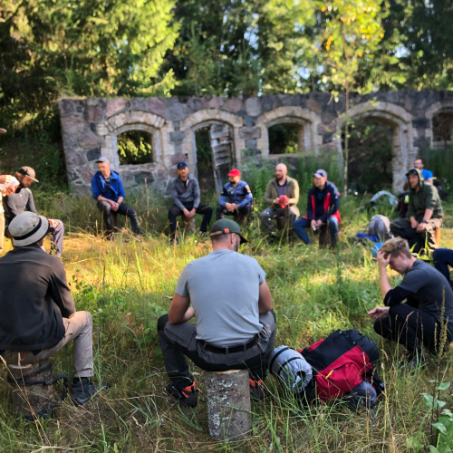 Grupa ar apmēram 20 vīrie&scaron;iem sēž aplī brīvā dabā pie vecas akmens ēkas drupām, kas atrodas meža ielokā. Daži vīrie&scaron;i sēž uz celmiem vai mugursomām, viens vīrietis stāv un runā, pārējie klausās. Apkārtne ir zaļa, ar zāli un kokiem, un notieko&scaron;ais izskatās kā sarunu vai atbalsta grupa dabā.