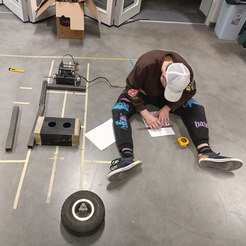 7.klases skolēns sēž darbnīcā uz grīdas un plāno sava elektroauto prototipu. Blakus uz grīdas redzami dažādi darba materiāli, riepa.