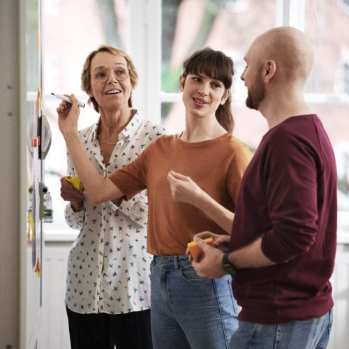 Divas sievietes un viens vīrietis stāv gai&scaron;ā telpā pie tāfeles un sarunājas.