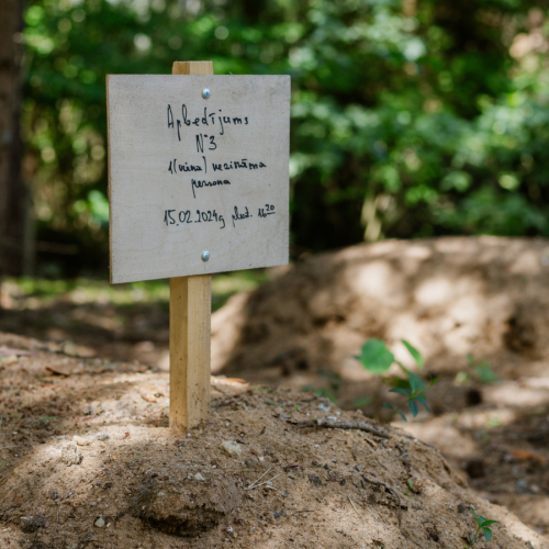 Pa&scaron;taisīta koka plāksne ar roku rakstītu uzrakstu, kas novietota uz smil&scaron;u kapa kopiņas. Uz plāksnes latvie&scaron;u valodā rakstīts: "Apbedījums Nr. 3, viena nezināma persona, 15.02.2024. plkst. 16.20". Fonā redzamas citas kapu kopiņas un zaļ&scaron; mežs.