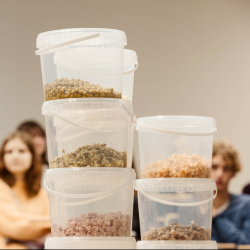 Plastmasas spaiņi, daļēji piepildīti ar dažādiem pārtikas produktiem, piemēram, gurķiem, kubiņos sagrieztu gaļu, kartupeļiem un citiem, sakrauti viens uz otra. Fonā redzami cilvēki, kas sēž un skatās uz priek&scaron;u izrādes laikā.