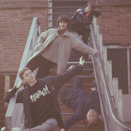 Četri jauni cilvēki pozē rado&scaron;ā un rotaļīgā veidā uz metāla kāpnēm pie ķieģeļu sienas. Viens no viņiem stāv ar roku uz margām, otrs atbalstījies pret margām apak&scaron;ā, tre&scaron;ais guļ otrādi pāri margām ar galvu uz leju, bet ceturtais sēž aug&scaron;pusē, atbalstījies pret margām. Attēls veidots siltos, retro toņos.