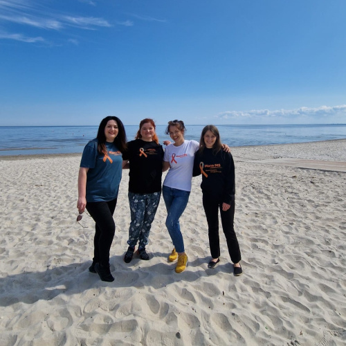 Četras sievietes stāv pludmalē un fonā redzama jūra un zilas debesis.