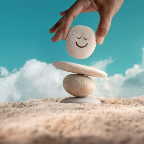 Attēlā redzama cilvēka roka, kas tur un novieto oļu ar uzgleznotu smaidīgu sejiņu uz citu, smil&scaron;ainā vidē sakrautu līdzsvarotu oļu kaudzītes. Fonā ir zilas debesis ar baltiem mākoņiem.