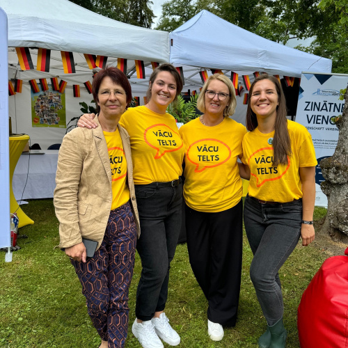 Attēlā redzamas četras smaido&scaron;as Vācijas vēstniecības Rīgā darbinieces, kuras tērpu&scaron;ās dzeltenos krekliņos ar uzrakstu &bdquo;vācu telts&ldquo;. Fonā redzama pati telts, kas izrotāta ar Vācijas karodziņiem un informatīviem attēliem par studiju iespējām Vācijā.