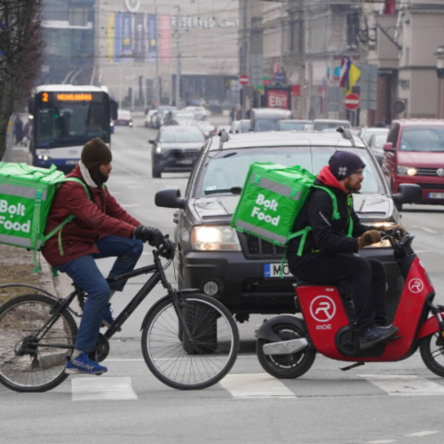 Attēlā redzams Rīgas ielas skats, kuru pa gājēju pārēju mēģina &scaron;ķērsot divi tum&scaron;as ādas krāsas vīrie&scaron;i: viens ar motorolleru, otrs ar riteni, abiem diviem zaļas Bolt ēdienu piegādes kastes uz muguras.
