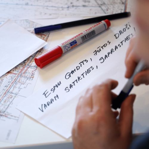 Uz galda dažādi papīri un rakstāmlietas, uz baltas lapas ar melnu flomāsteru uzrakstīts "Esmu gaidīts, jūtos dro&scaron;i, varam satikties, saprasties".