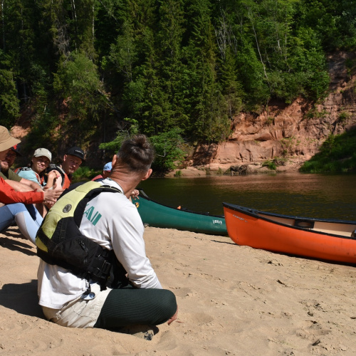 Grupa cilvēku sēž smil&scaron;ainā upes krastā pie kanoe laivām, baudot saulainu dienu dabā. Priek&scaron;plānā redzams vīrietis ar glāb&scaron;anas vesti, bet fonā &ndash; pārējie dalībnieki sarunājas un smaida. Aiz viņiem plūst upe ar kanoe laivām krastā, un otrā krastā paceļas sarkanas klintis un blīvs zaļ&scaron; mežs. Attēls atspoguļo aktīvu atpūtu pie dabas un draudzīgu kopā bū&scaron;anu.