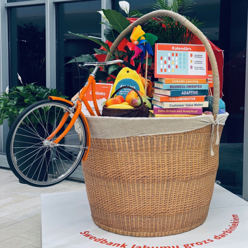 Ofisa vidē novietots liels pīts grozs, no kura ārā redzama reāla velosipēda priek&scaron;ējā riepa ar stūri, groza virspusē redzamas palielināta izmēra grāmatas, dažādi augļi un citi priek&scaron;meti