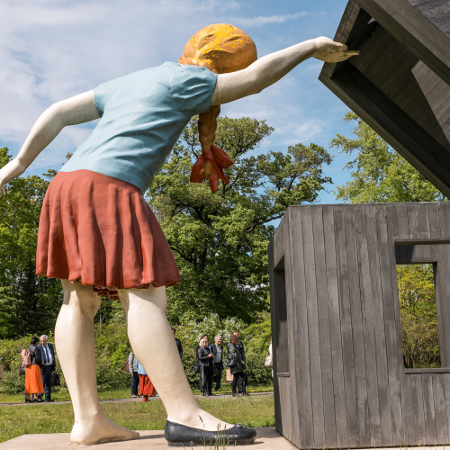 Attēlā redzams liels vides, mākslas objekts - stilizēta meitene ģērbta īsos bruņčos un krekliņā kura ver vaļā jumtu blakus eso&scaron;ajai, stilizētai koka ēkai. Fonā zilas debesis, zaļi koki un cilvēki.