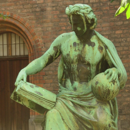 Attēlā redzama skulptūra, aiz tās sarkanu ķieģeļu siena.