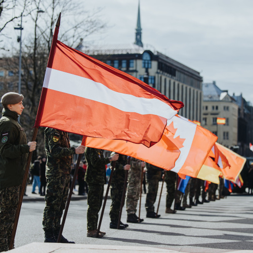 Brīvības pieminekļa laukums, rindā stāv karavīri, katrs rokā tur karogu, priek&scaron;plānā Latvijas karogs, tad Kanādas karogs, pēc tam citi karogi, kas nav iz&scaron;ķirami.