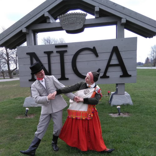 Zaļā pļavā priek&scaron;plānā redzams dejotāju pāris, kas stāv viens otram līdzās, sadevu&scaron;ies rokās krusteniskajā satvērienā, atlieku&scaron;ies viens no otra uz t.s. ārējās kājas pusi, bet skatās viens otram acīs. Puisim gai&scaron;i pelēks vadmalas tērps ar baltu kreklu un sarkanu lakatiņu ap kaklu. Pui&scaron;a kamzolim (t.s. svārkiem) melnu diegu iz&scaron;uvumi uz krūtīm gar aizdari un gar piedurknēm, galvā melns cilindrs un kājās melni zābaki. Partnerei sūnu zaļā sievas cepure, sūnu zaļa jaka ar tum&scaron;i zaļā samta apmalēm, Nīcas oranži-sarkanie brunči un baltā villaine, kas gar malu iz&scaron;ūta ar krāsainu diegu rakstiem un sasprausta ar saktām uz kreiso plecu. Aiz cilvēkiem fonā zem divslīpu jumtiņa uz pelēka pamata ar lieliem melniem burtiem uzraksts - NĪCA.