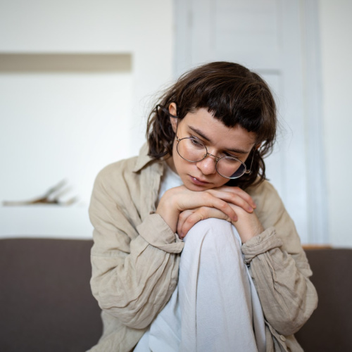 Jauna persona ar brūniem matiem un brillēm sēž uz dīvāna, apskāvusi ceļgalus un skatās uz leju, izskatoties domīga vai noskumusi.