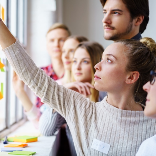 Jauna sieviete ar vārdu zīmīti 'Emily' pie loga līmē krāsainu līmlapiņu, kamēr aiz viņas uzmanīgi vēro vēl pieci cilvēki &mdash; diskusijā vai darbnīcā iesaistīta komanda, kas strādā pie ideju ģenerē&scaron;anas.