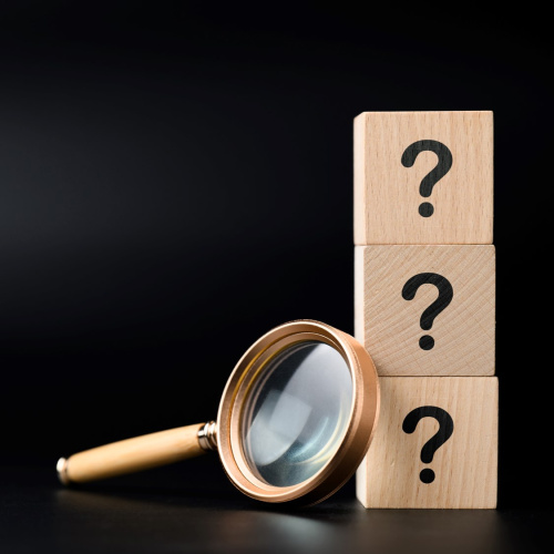 Uz melna fona trīs koka klucī&scaron;i ar jautājuma zīmēm un lupa.