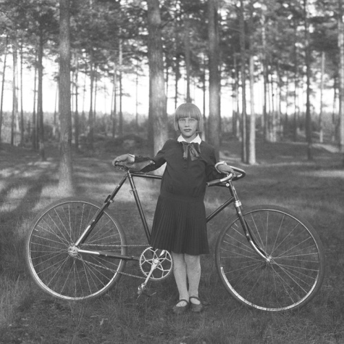 Sena fotogrāfija, kurā redzama meitene, kas pozē pie velosipēda, aizmugurē mežs.