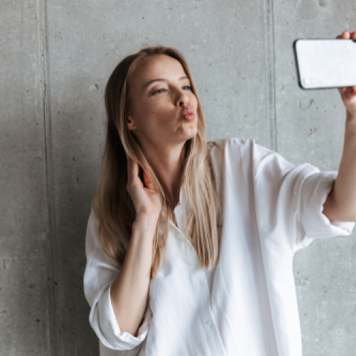 Jauna sieviete gai&scaron;ā, brīvā kreklā uz pelēka fona uzņem pa&scaron;bildi jeb selfiju ar viedtālruni, viegli sūtot gaisa skūpstu kamerai.