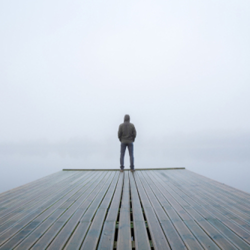 Cilvēks ar kapuci mugurā stāv uz koka laipas, kas iestiepjas klusā un miglainā ūdens virsmā, skatoties tālumā. Apkārtne ir miglā tīta un rada mierīgu un noslēpumainu atmosfēru.