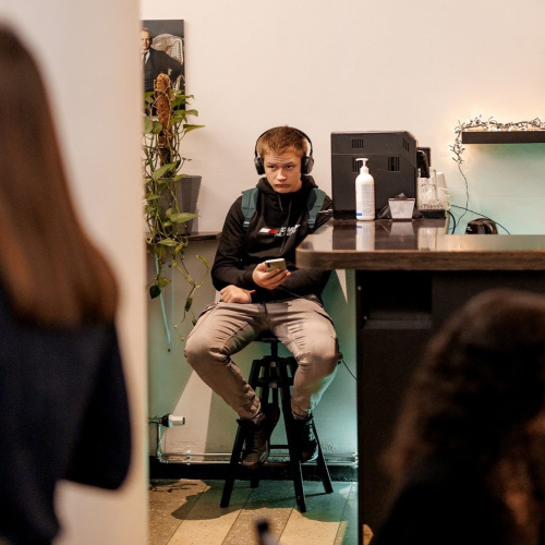 Jauns cilvēks ar austiņām sēž uz augsta krēsla pie letes, turot rokās viedtālruni. Aiz viņa redzama kafijas automāta iekārta, dezinfekcijas līdzekļa pudele un plaukts ar gaismas virteni. Fonā vairākas personas, daļēji redzamas, iesaistītas sarunās vai kustībā.