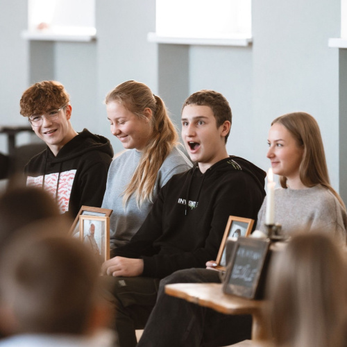 Četri pusaudži sēž rindā un izskatās pārsteigti par kaut ko ārpus kadra.