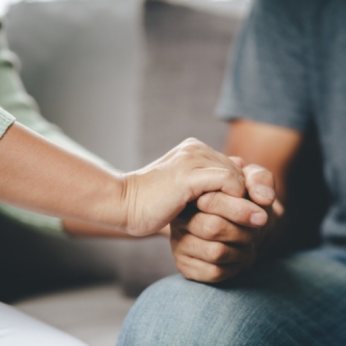 Divu cilvēku tuvplāns, kur viens cilvēks mierino&scaron;i tur otra roku, simbolizējot atbalstu, līdzjūtību vai rūpes. Fonā ir mierīgi, neitrāli toņi.