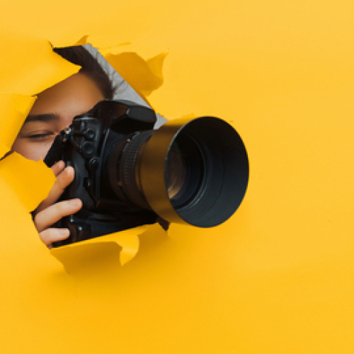 Cilvēks, kur&scaron; caur dzeltenā papīra sienā izplēstu caurumu fotografē ar profesionālu fotokameru. Fonā spilgti dzeltens papīrs, kas rada kontrastainu un uzmanību piesaisto&scaron;u efektu.