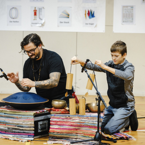 Vīrietis ar tetovējumiem un zēns spēlē uz dažādiem perkusijas instrumentiem uz grīdas.