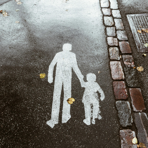 Lietainā laikā uz asfalta baltā krāsā uzzīmēts pieaugu&scaron;ā un bērna simbols, kas iet roku rokā.