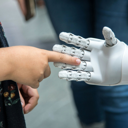 Bērns pieskaras ar pirkstu robota rokai, simbolizējot cilvēka un tehnoloģiju mijiedarbību.