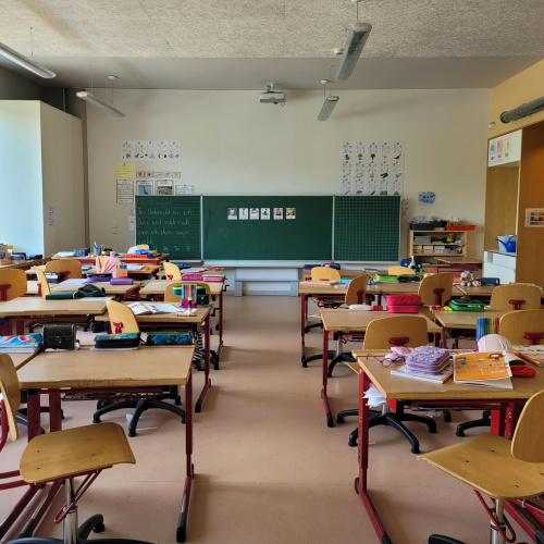 Skolas klase, kurā ir rindas ar skolēnu galdiem un krēsliem, kas vērsti pret zaļu tāfeli sienas priek&scaron;ā. Uz galdiem ir dažādi mācību materiāli. Klase izskatās tuk&scaron;a un gatava skolēnu iera&scaron;anās.