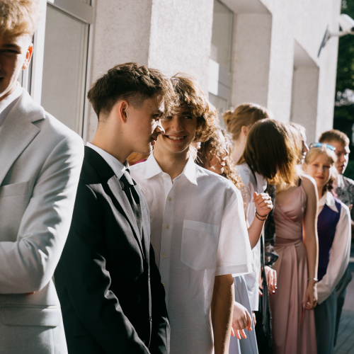 Grupa jaunie&scaron;u, ģērbu&scaron;ies svinīgos apģērbos, stāv rindā pie skolas.