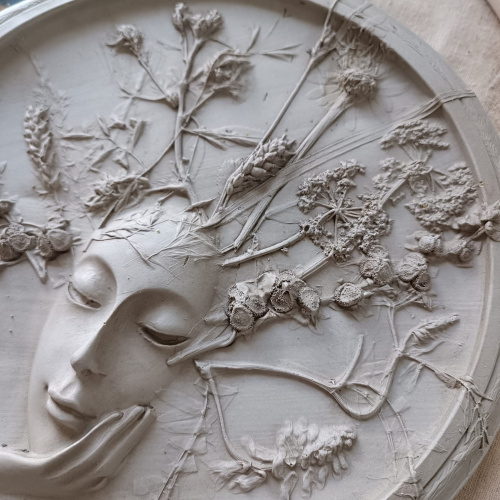 Botāniskā bareljefa detaļa, kurā attēloti sievie&scaron;u seja, augi, lapas un ziedi.