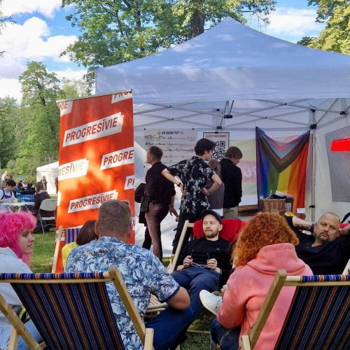 Aplī uz pludmales krēsliem sēz dažādi cilvēki. Fonā balta telts un sarkans baneris ar uzrakstu "Progresīvie".