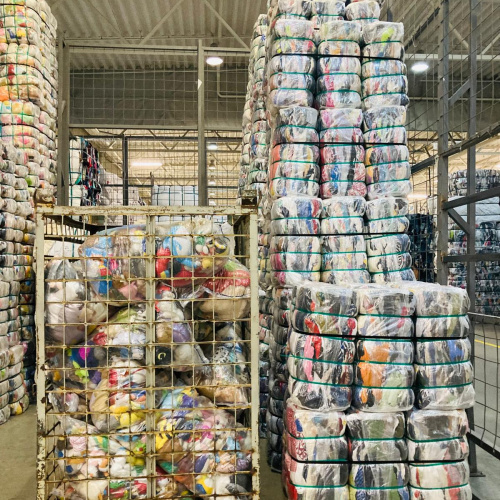 Noliktava ar sakrautiem, plastmasā iepakotiem lietoto apģērbu saiņiem un metāla konteineru, kas piepildīts ar krāsainiem tekstilizstrādājumiem.