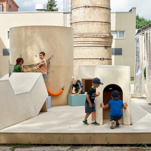 Bērni rotaļājas uz mūsdienīga koka rotaļu laukuma ar ģeometriskas formas konstrukcijām. Fonā atrodas ķieģeļu skurstenis un mūsdienīgas ēkas.