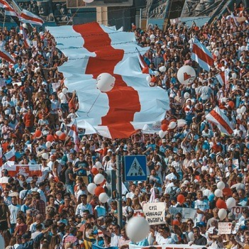 Liela cilvēku demonstrācija ar vēsturiskajiem Baltkrievijas karogiem, plakātiem un baloniem, pūlis piepilda ielu, izrādot politisku atbalstu un vienotību.
