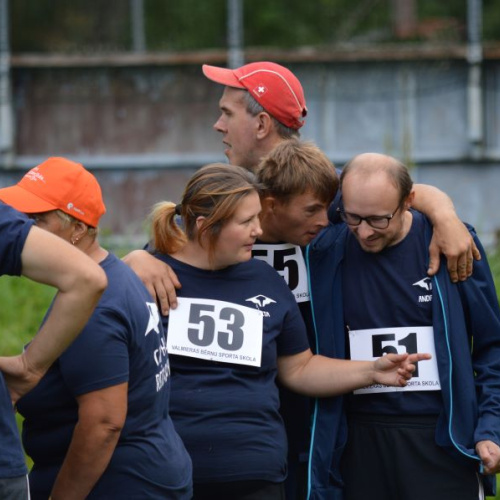 Cilvēku grupa sporta sacensību laikā, visi ģērbu&scaron;ies vienādos sporta kreklos ar numuriem, cits citu atbalsta un apskauj, izrādot komandas garu un draudzību.