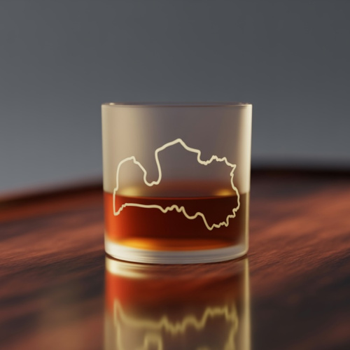 Uz koka galda alkohola glāze ar Latvijas kontūru un brūnu dzērienu tajā.