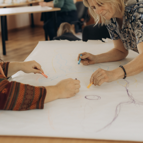 Attēlā redzamas divu sievie&scaron;u rokas uz baltas papīra lapas, kuras zīmē ar krāsainiem krītiņiem