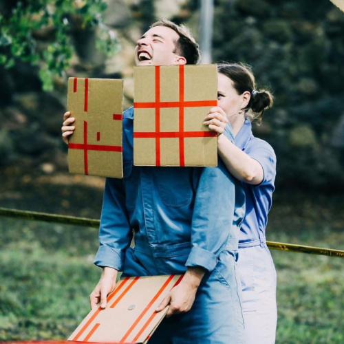 Attēlā redzami divi cilvēki brīvā dabā. Sieviete stāv tie&scaron;i aiz vīrietim aiz muguras un viņa priek&scaron;ā tur divus kartonus ar sarkanām svītrām, kas veido krustus. Vīrietis smejas.