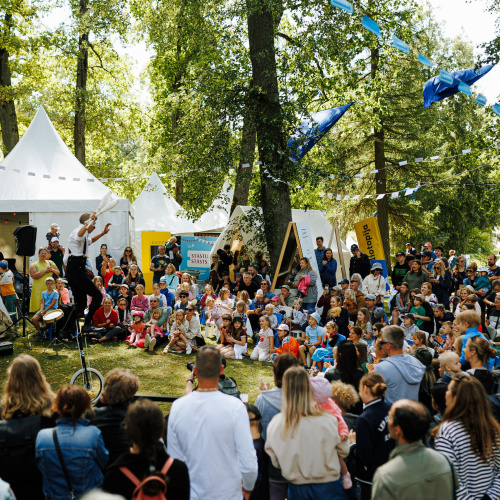 Cilvēku grupa piedalās āra pasākumā parkā, kur mākslinieks uz vienriteņa žonglē auditorijas priek&scaron;ā. Apkārt viņam sēž un stāv skatītāji, tostarp bērni un pieaugu&scaron;ie, baudot priek&scaron;nesumu. Baltas teltis un izkārtie karodziņi veido pasākuma atmosfēru.